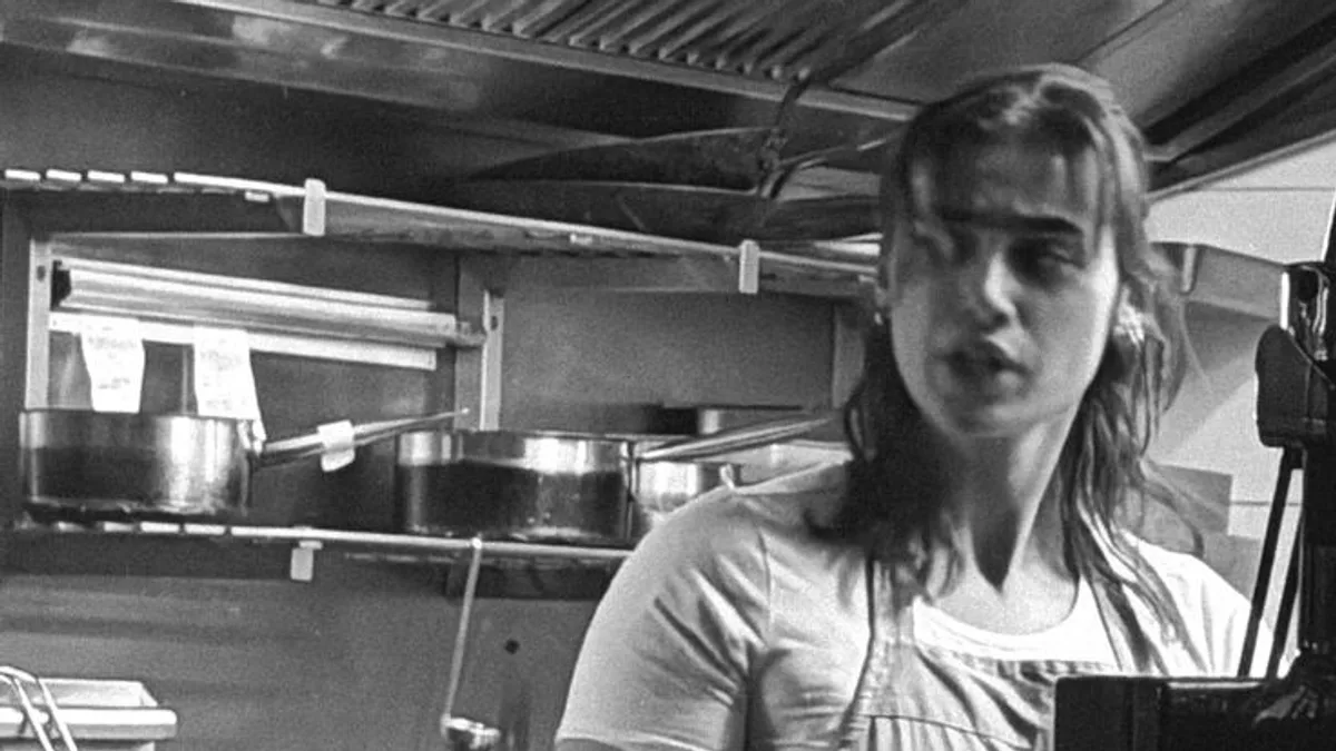 A restaurant chef standing with arms crossed at the line in a stainless commercial kitchen, sauce pots on the shelf behind her, pausing before service