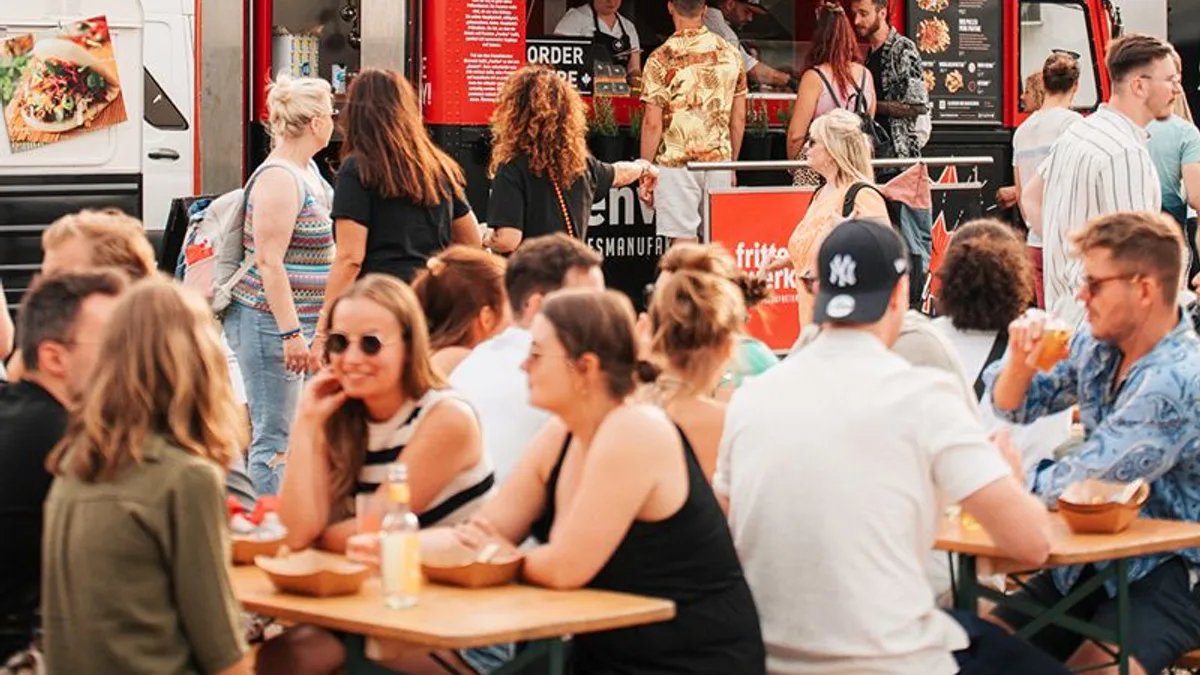 A busy red food truck serving a crowd seated at outdoor picnic tables on a sunny day
