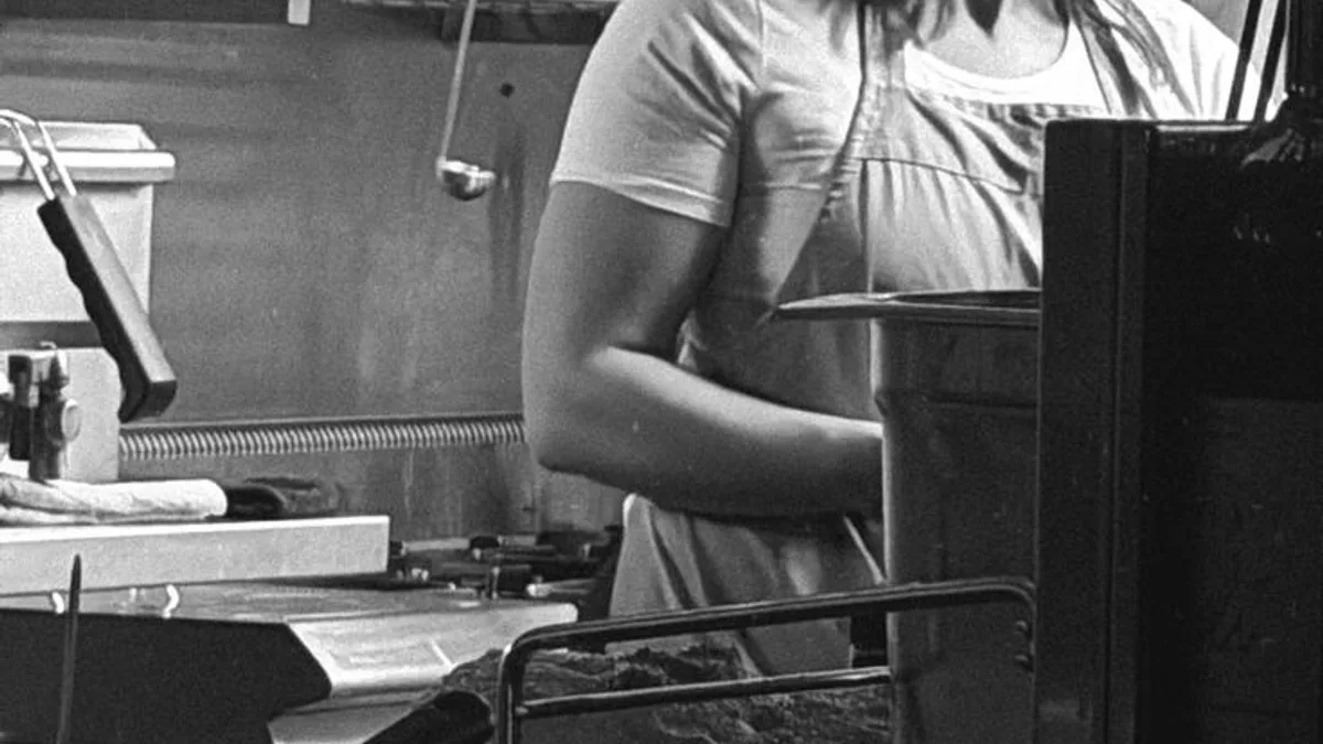 Black-and-white photograph of a line cook at her station beneath a stainless exhaust hood in a commercial kitchen