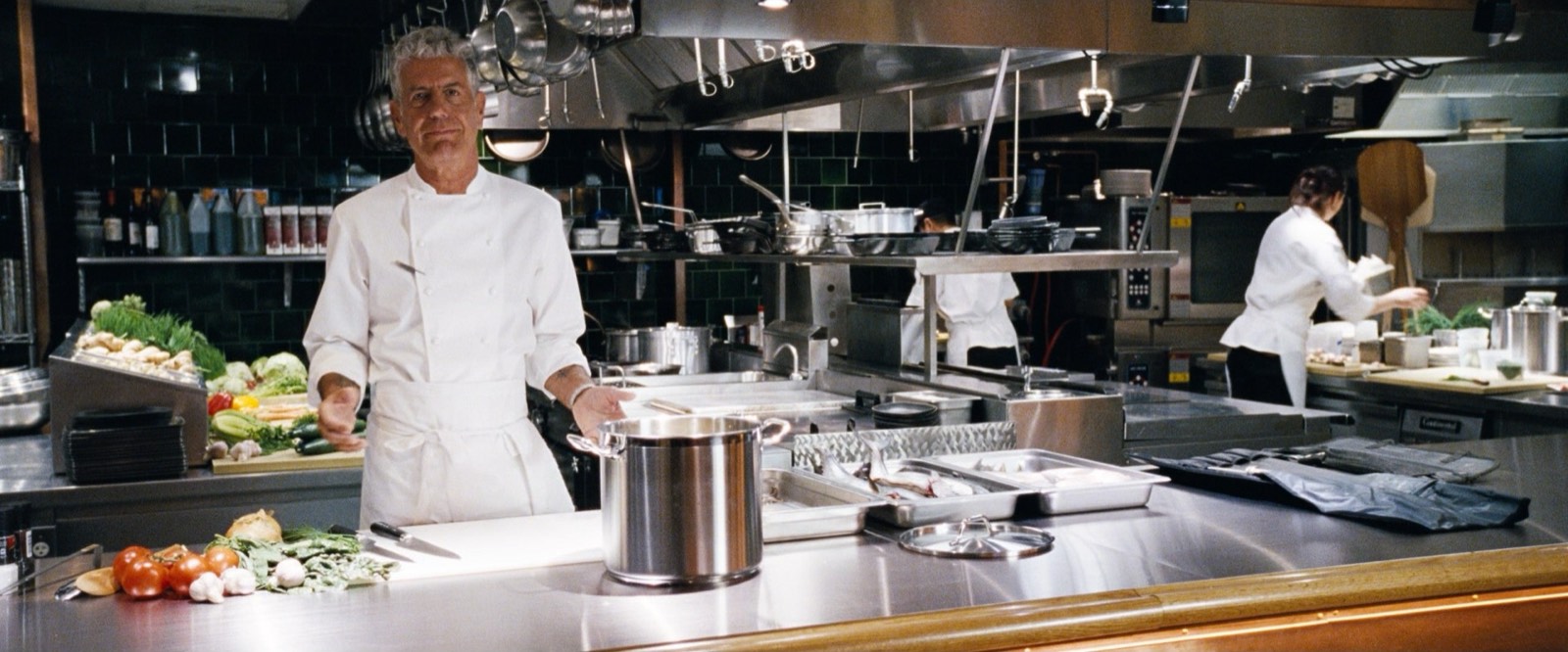 A chef standing in a professional kitchen with fresh ingredients laid out for service.
