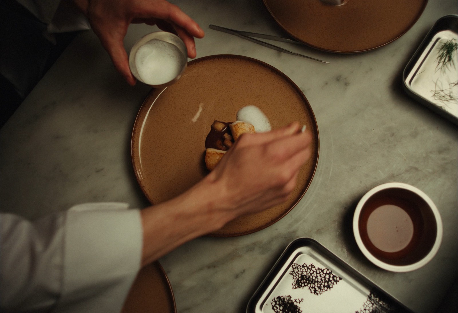 A chef plating a fine-dining dish with tweezers on a ceramic plate.